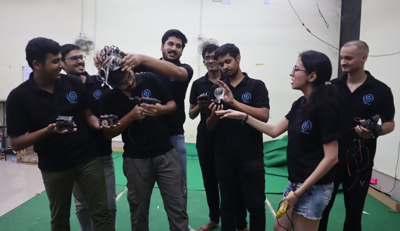 KRSSG teammates laughing and goofing around while holding robot parts, with one robot balanced on a teammate’s head.