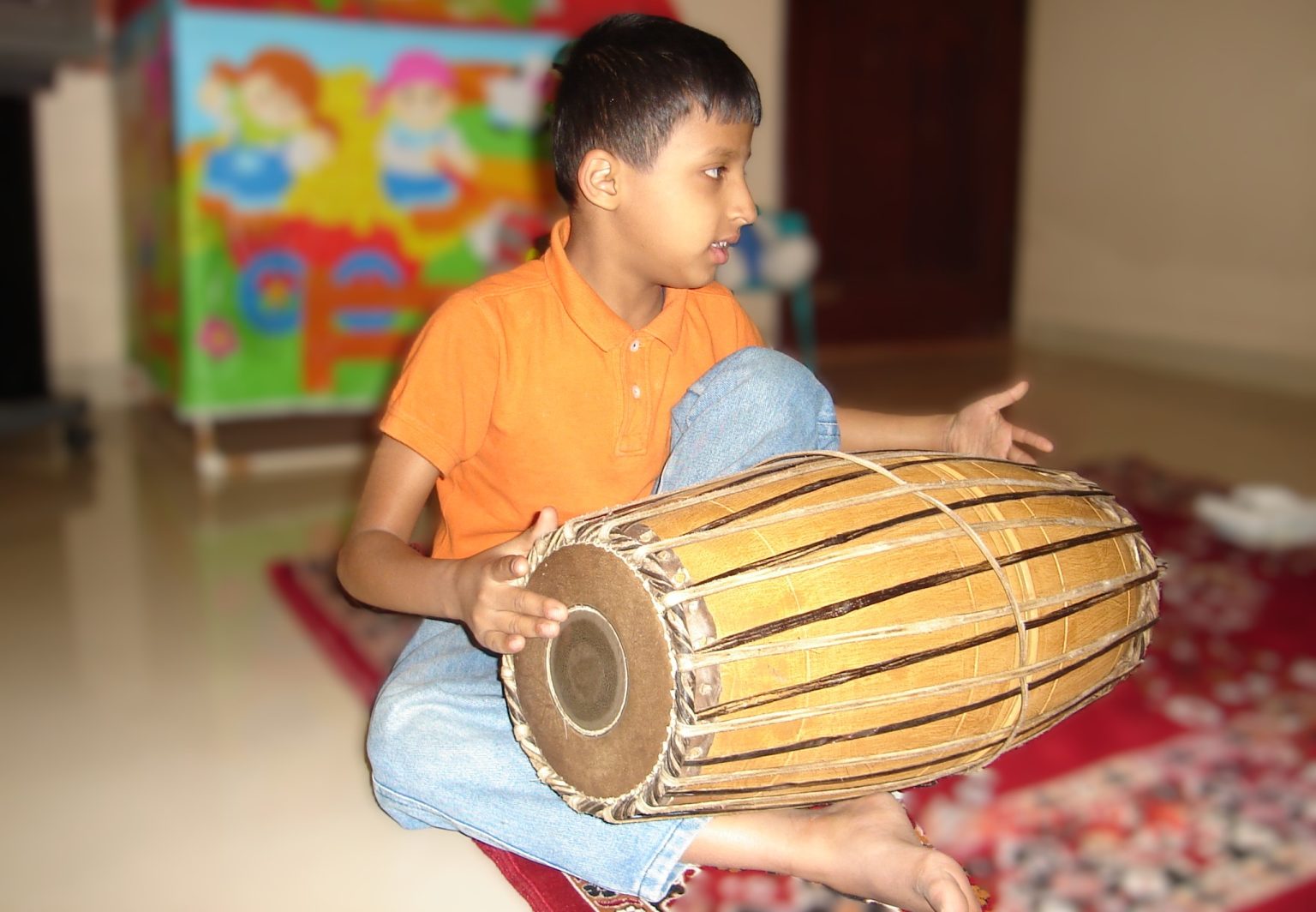 Me as a child, seated with a Mridangam across my lap, wearing an orange shirt.
